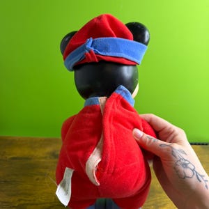 May include: A plush toy featuring a black head with large ears, wearing a red hat with a blue band and a red jumpsuit with blue accents. The toy is held by a hand with a floral tattoo.