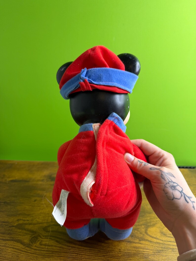 May include: A plush toy featuring a black head with large ears, wearing a red hat with a blue band and a red jumpsuit with blue accents. The toy is held by a hand with a floral tattoo.