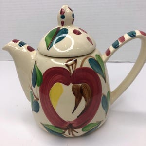 Puede incluir: Una tetera de cerámica blanca con un diseño de manzana roja. La tetera tiene una tapa con una hoja verde y lunares rojos. El asa está rayada de rojo, blanco y azul.