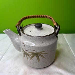 Puede incluir: Una tetera de cerámica moteada con un diseño de hojas de bambú. La tetera tiene una tapa con un pomo oscuro y un asa tejida. El pico está en el lado izquierdo. El fondo es verde liso.