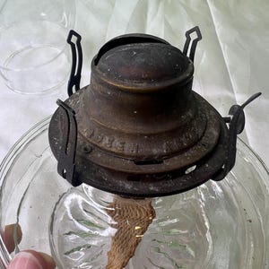 Puede incluir: Una l&aacute;mpara de aceite antigua con una base de vidrio transparente y un quemador de lat&oacute;n deslustrado. El quemador tiene una mecha y est&aacute; unido a un marco de metal. La l&aacute;mpara est&aacute; dise&ntilde;ada para contener aceite y proporcionar luz. La base de vidrio tiene un patr&oacute;n decorativo.