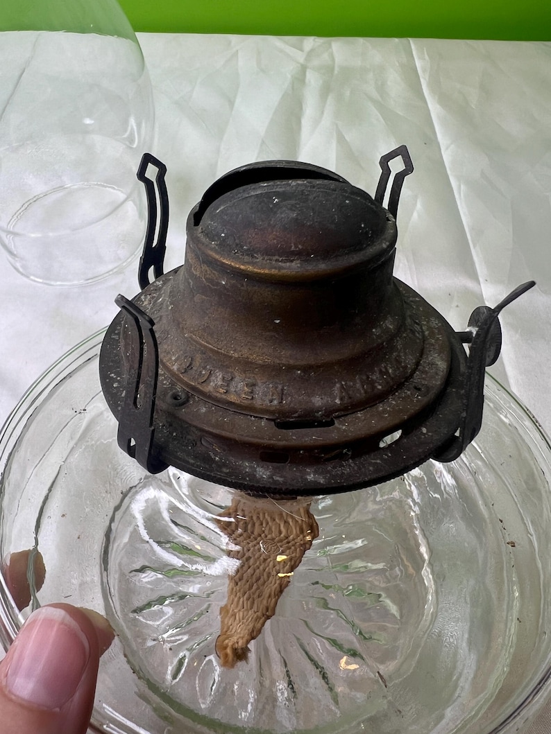 Puede incluir: Una l&aacute;mpara de aceite antigua con una base de vidrio transparente y un quemador de lat&oacute;n deslustrado. El quemador tiene una mecha y est&aacute; unido a un marco de metal. La l&aacute;mpara est&aacute; dise&ntilde;ada para contener aceite y proporcionar luz. La base de vidrio tiene un patr&oacute;n decorativo.