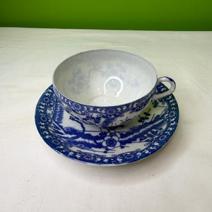 May include: A white teacup and saucer with blue floral patterns. The cup has a handle and sits on a matching saucer. The blue design includes flowers and foliage, creating a classic, elegant look. The set is likely porcelain or ceramic.