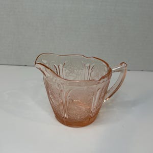 Könnte beinhalten: Eine rosa gläserne Sahnegießer mit einem Blumenmuster. Der Sahnegießer hat einen Griff und einen gewellten Rand.
