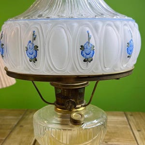 Könnte beinhalten: Vintage Öllampe mit einem klaren Glassockel und einem Schirm aus Milchglas. Der Schirm ist mit blauen Blumenmustern und einer blauen Bordüre verziert. Die Lampe hat einen messingfarbenen Metallrahmen.