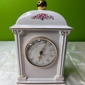 May include: White ceramic quartz clock with a gold-colored bezel and Roman numerals. The clock features a floral design and a gold-colored top. The clock is rectangular with a curved top and decorative columns on each side.