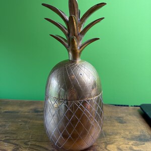 May include: A vintage brass pineapple-shaped container with a removable lid. The pineapple has a textured surface with a diamond pattern on the body and detailed leaves at the top. The container is likely used for storage or decoration.