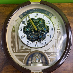 May include: A Seiko Melodies in Motion wall clock with a dark brown oval frame. The clock face features a white dial with black numerals, a central dark blue rotating gear mechanism with blue crystals, and a gold-toned city skyline illustration.