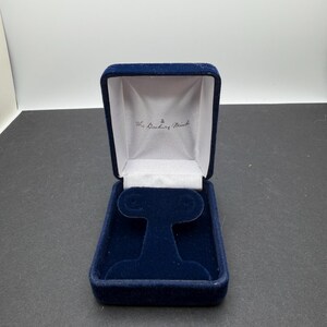 May include: A navy blue velvet jewelry box with a hinged lid. The interior features a white satin lining with the text The Danbury Mint printed in black, and a navy blue velvet insert designed to hold a pair of earrings in an upright position.