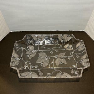 May include: A clear glass serving tray with a floral design etched into the surface. The tray has a rectangular shape with slightly rounded corners.