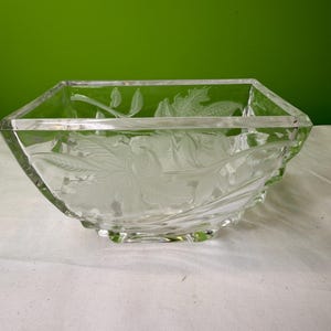 以下が含まれることがあります： 透明な正方形のガラスボウル。エッチングされた花の模様が施されています。ボウルの縁はわずかに波状で、小さな正方形の台座の上に置かれています。花の模様はボウルの側面に施されており、装飾的なアクセントになっています。