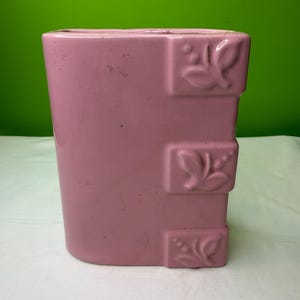 Könnte beinhalten: Eine rechteckige, rosa Keramikvase mit leicht gebogenem Boden. Die Vase hat drei rechteckige, erhabene Dekorationselemente mit floralem Design. Der Hintergrund ist einfarbig grün.