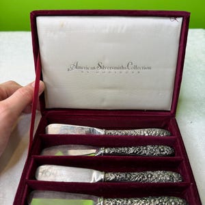 May include: A set of four silver butter knives with decorative floral handles, displayed in an open burgundy velvet-lined box. The box features the text "American Silversmiths Collection by Goddinger" on the inside lid.