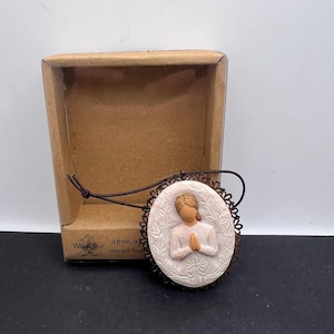 Könnte beinhalten: Ein ovales, cremefarbenes Anhänger mit einem braunen, verzierten Rand. Der Anhänger zeigt eine geschnitzte Figur mit gefalteten Händen im Gebet. Der Anhänger ist an einer braunen Kordel befestigt und liegt neben einer braunen Pappschachtel.