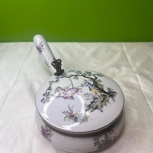Può includere: Scatola portagioie in porcellana bianca con coperchio raffigurante una scena pastorale con figure e decorazioni floreali. La scatola ha un manico e un bordo in metallo argentato. Il manico è decorato con motivi floreali.