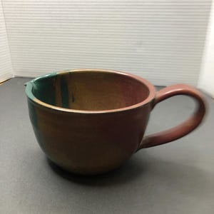 Może przedstawiać: Brązowy kubek ceramiczny z zielono-brązową glazurą. Kubek ma zaokrąglony kształt i uchwyt.