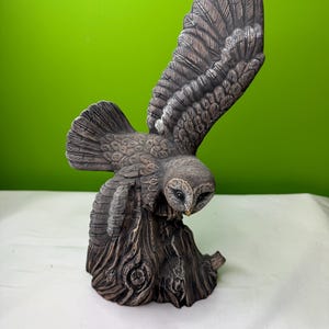 Może przedstawiać: Szczegółowa ceramiczna figurka sowy z rozłożonymi skrzydłami, siedząca na teksturowanym pniu drzewa. Sowa jest głównie szara i brązowa, ze skomplikowanymi detalami piór. Tło jest jasnozielone.