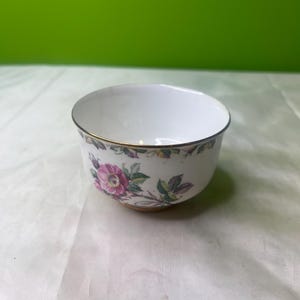May include: White ceramic bowl with a floral design featuring pink roses and green leaves. The bowl has a gold rim and base. The floral pattern wraps around the exterior of the bowl, adding a touch of elegance.