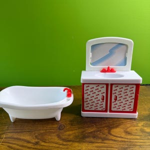 Puede incluir: Un juego de bañera y tocador blancos en miniatura. El tocador tiene un espejo, un grifo rojo y un armario rojo y blanco. La bañera tiene un grifo rojo. El armario del tocador tiene el texto "CPK" repetido.