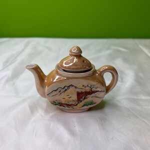 May include: A small, tan ceramic teapot with a lid. The teapot features a painted landscape scene with mountains, trees, and a red structure. The spout and handle are curved, and the lid has a small knob. The teapot is likely vintage.