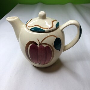 May include: A white ceramic teapot with a brown, green, and burgundy apple design. The teapot has a lid and a handle.