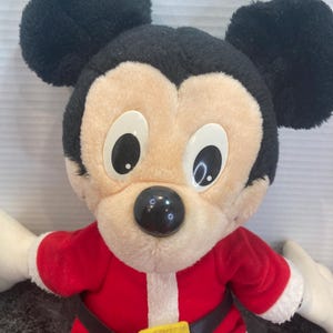 May include: A plush Mickey Mouse toy wearing a red Santa Claus outfit with white trim, a black belt, and a yellow buckle. The character has large black ears, a beige face, white plastic eyes, and a round black nose, presented in a festive style.