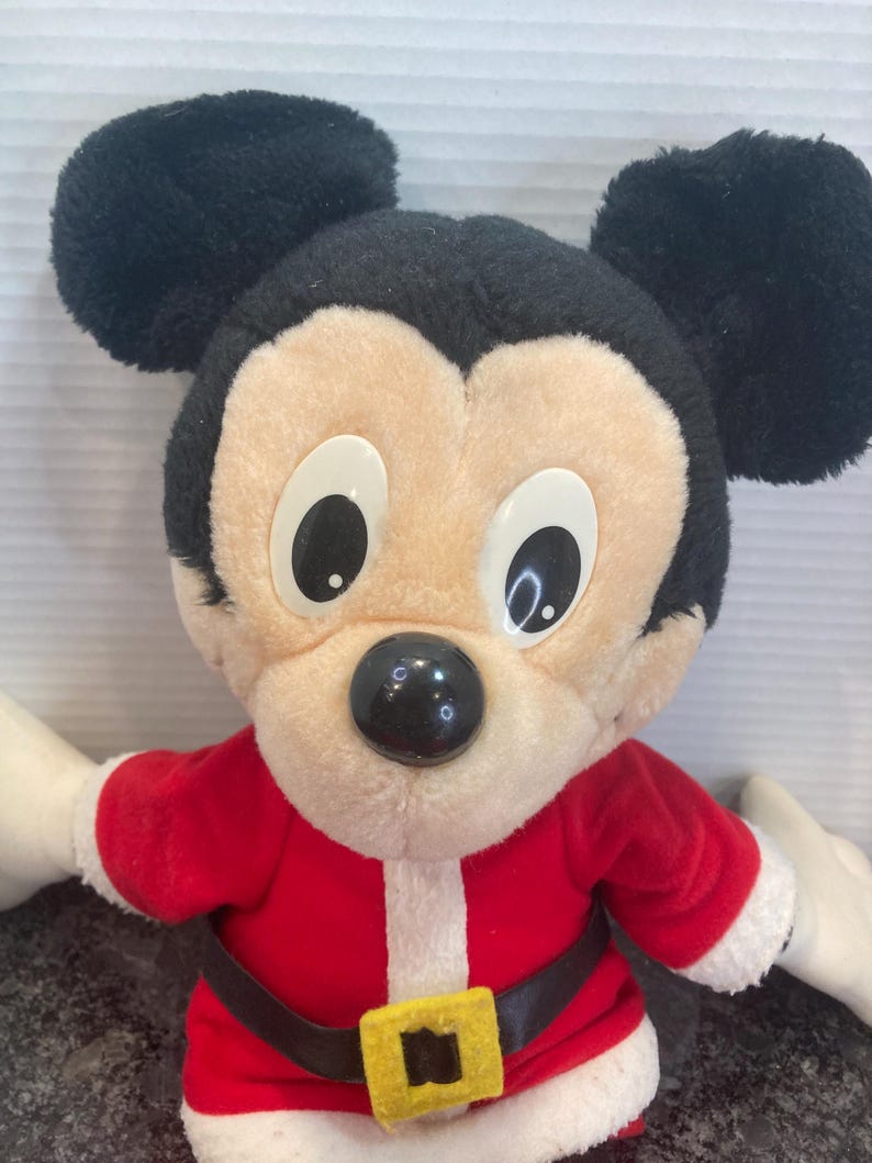 May include: A plush Mickey Mouse toy wearing a red Santa Claus outfit with white trim, a black belt, and a yellow buckle. The character has large black ears, a beige face, white plastic eyes, and a round black nose, presented in a festive style.