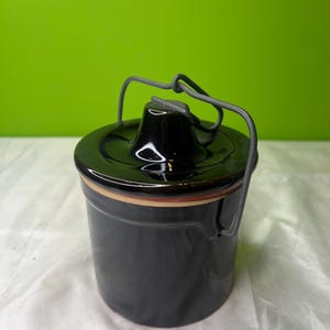 May include: A black ceramic jar with a lid and a metal handle. The jar is cylindrical with a brown and beige band around the middle. The lid has a small, raised centre. The handle is made of grey wire.
