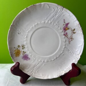May include: White porcelain saucer with a floral design in shades of pink, yellow, and green. The saucer has a raised, embossed pattern around the edge.