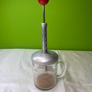 Pode incluir: Uma ferramenta de cozinha vintage com um copo medidor de vidro transparente, parte superior de metal e uma alça de bola vermelha. O copo tem marcações de medição. A ferramenta foi projetada para misturar ou mexer ingredientes. O fundo é verde liso.