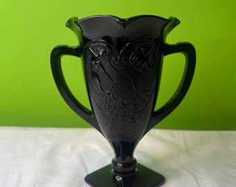 Peut inclure: Vase en verre noir avec deux anses et un pied carré. Le vase est orné d'un motif gravé de deux figures et d'un cœur.