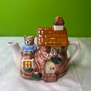 Puede incluir: Una tetera de cerámica caprichosa con forma de casa en el árbol, con tejado marrón y chimenea. Se ven dos osos de peluche, uno en el porche y otro en una ventana. La tetera tiene un pico y un asa blancos, con detalles de puerta y ventana.
