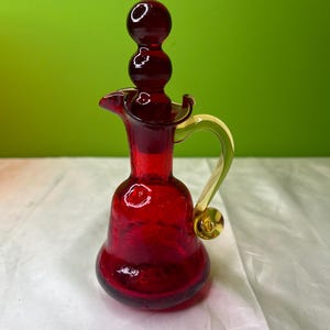 May include: A small, ruby red glass pitcher with a spherical base and a long, slender neck. The handle is a pale yellow-green color, and the stopper is a series of three stacked spheres. The pitcher has a spout and a decorative design.