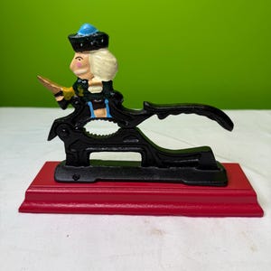 Puede incluir: Un cascanueces vintage de hierro fundido con forma de soldado. El soldado tiene un sombrero azul, pelo blanco y una espada dorada. El cascanueces negro está montado sobre una base roja. La base mide unos 15 cm de largo.