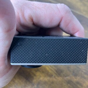 May include: A black action camera with a textured, diamond-patterned surface. The camera has a silver trim along the bottom edge and a small, black button on the side. The camera is held in a hand.