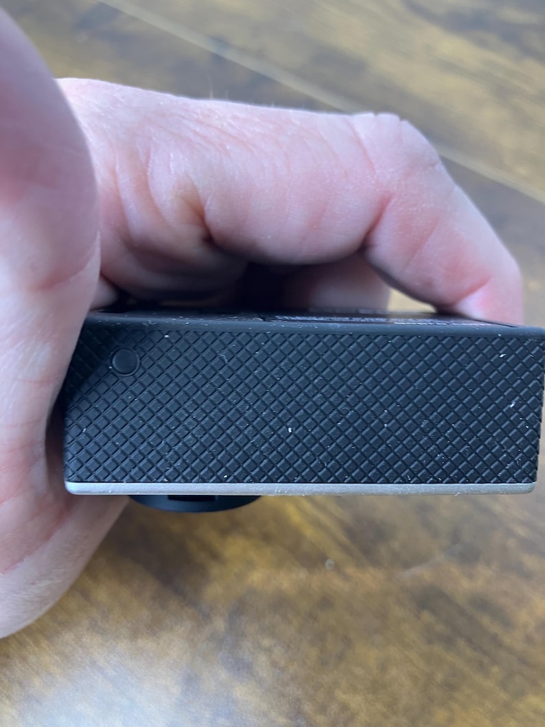 May include: A black action camera with a textured, diamond-patterned surface. The camera has a silver trim along the bottom edge and a small, black button on the side. The camera is held in a hand.