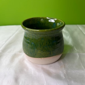 Può includere: Un piccolo vaso in ceramica verde con un bordo bianco. Il vaso ha una smaltatura verde macchiata con accenti blu.