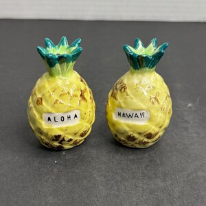 Puede incluir: Un juego de dos saleros y pimenteros de cerámica con forma de piña. Los saleros son amarillos con detalles marrones y tapas verdes. Un salero dice "ALOHA" y el otro dice "HAWAII".