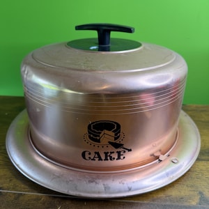 Peut inclure: Un présentoir à gâteau de couleur cuivre avec une poignée noire et une base ronde. Le côté du présentoir porte le mot "CAKE" et un dessin de gâteau avec une spatule.