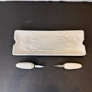 Puede incluir: Plato de servir de cerámica blanca con un diseño en relieve de una mazorca de maíz. Dos pequeños cuchillos de mantequilla de cerámica blanca con forma de mazorca de maíz con hojas plateadas.