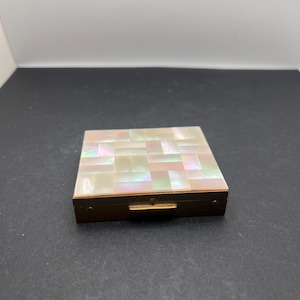 Könnte beinhalten: Ein quadratischer, antiker Kompaktpuder mit einer Deckplatte aus Perlmuttmosaik. Die schillernden Fliesen sind in einem rechteckigen Muster angeordnet. Der Kompaktpuder hat einen goldfarbenen Metallrahmen und einen kleinen Verschluss.