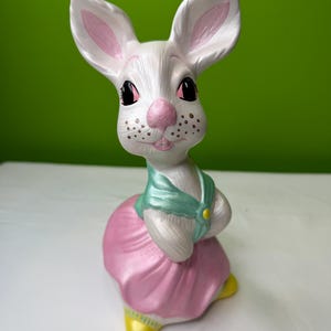 May include: A ceramic Easter bunny figurine. The bunny is white with pink inner ears, a pink nose, and black eyes. It wears a pink dress, a green shawl, and yellow shoes. The bunny has a smiling expression.