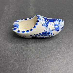 May include: A small, white ceramic Dutch shoe with blue and white Delftware designs. The shoe has a windmill and other traditional Dutch imagery painted on it.