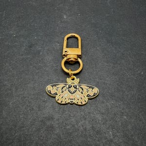 May include: A gold-toned metal keychain featuring a moth-shaped charm. The moth charm is decorated with black, cream, and soft pink enamel fill, showcasing intricate wing patterns with a central star detail and a gold-colored lobster clasp.