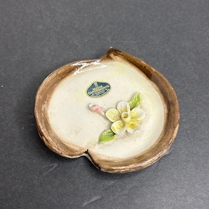 May include: A small, white ceramic dish with a brown rim and a raised flower design. The dish has a label that reads "NUOVA CAPODIMONTE CERAMICA ARTISTICA"