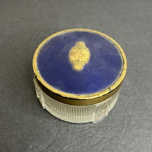 Può includere: Un barattolo di vetro vintage con un coperchio blu e oro. Il coperchio ha un motivo decorativo dorato al centro.