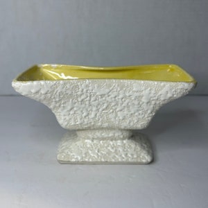 May include: A white ceramic planter with a yellow glaze inside. The planter has a textured surface and a square base.