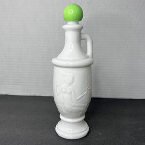 May include: A white ceramic bottle with a green stopper. The bottle has a handle and a decorative relief of a woman. The bottle is approximately 7 inches tall.