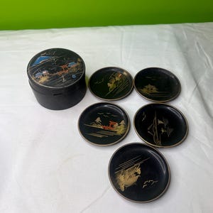 May include: A set of black coasters with gold and red Japanese-style landscape designs. The set includes a matching round box with a similar design on the lid. The coasters and box are likely made of a lacquered material.