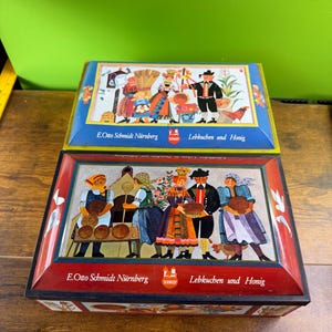 May include: Two vintage metal boxes with colourful folk art scenes. The boxes feature illustrations of people in traditional clothing, with text that reads "E. Otto Schmidt Nürnberg" and "Lebkuchen und Honig". The boxes are blue and red.